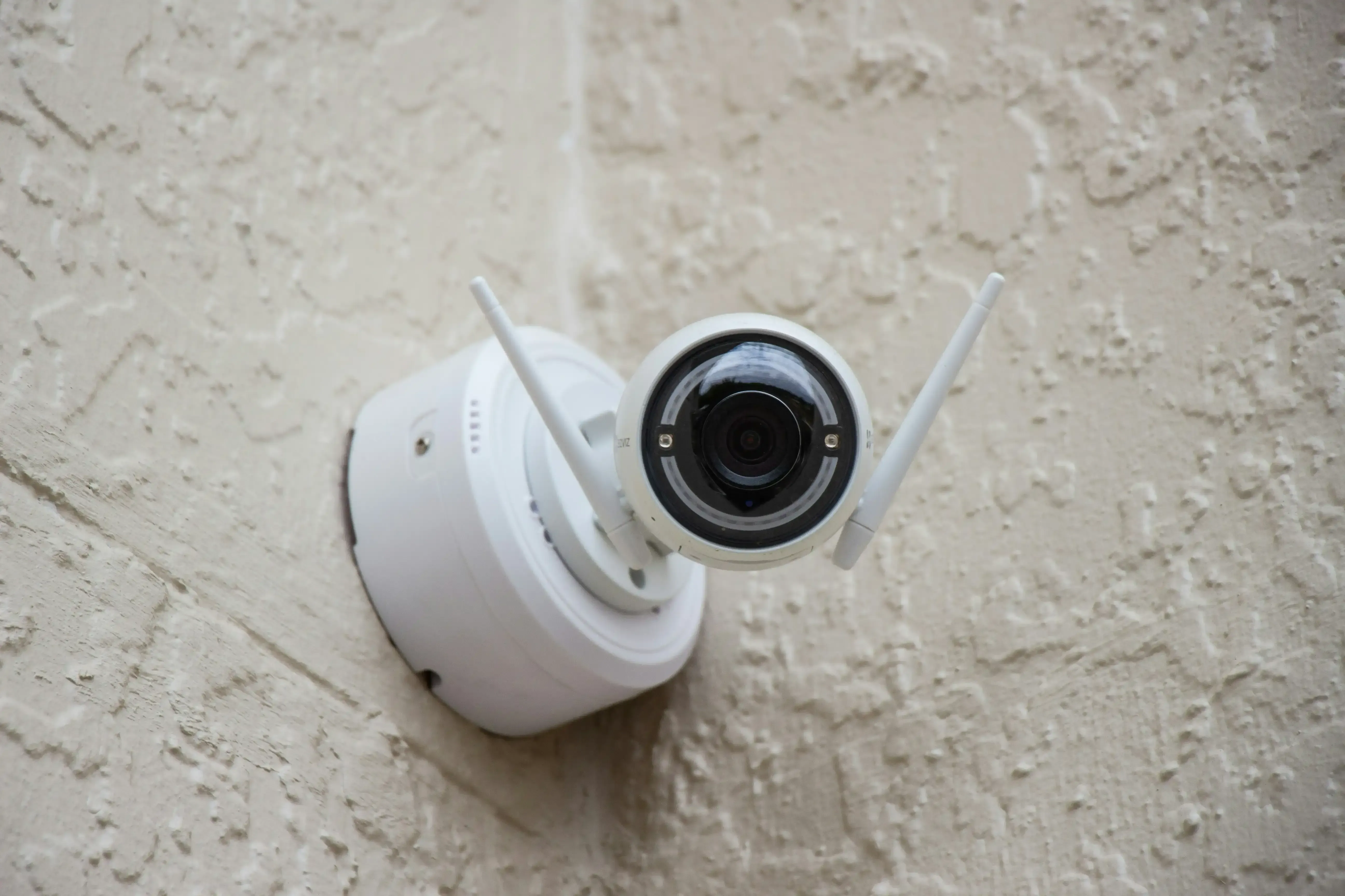 White security camera mounted on a textured beige wall, with two antennas extending outward.